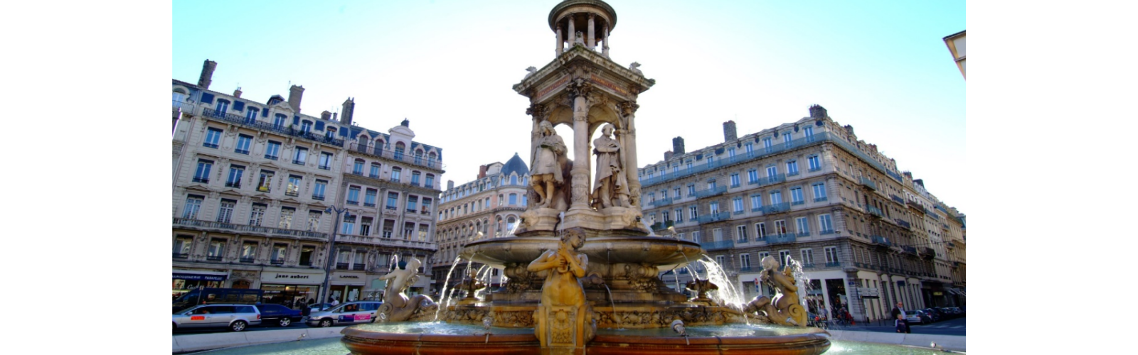 An old fountain in a city square of Lyon. The fountain has ornate detailing and is surrounded by shops and apartments.